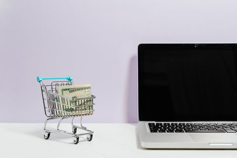 Laptop next to miniature shopping cart filled with money